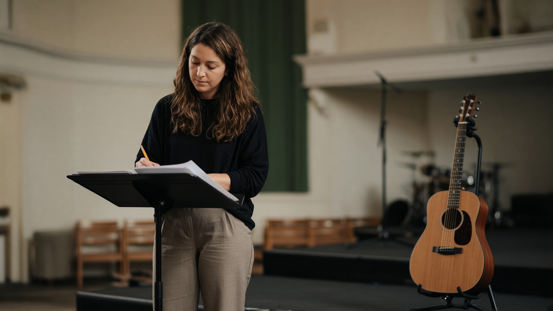 A worship leader at a music stand before a service, marking keys on a printed set list with the stage still dark behind her